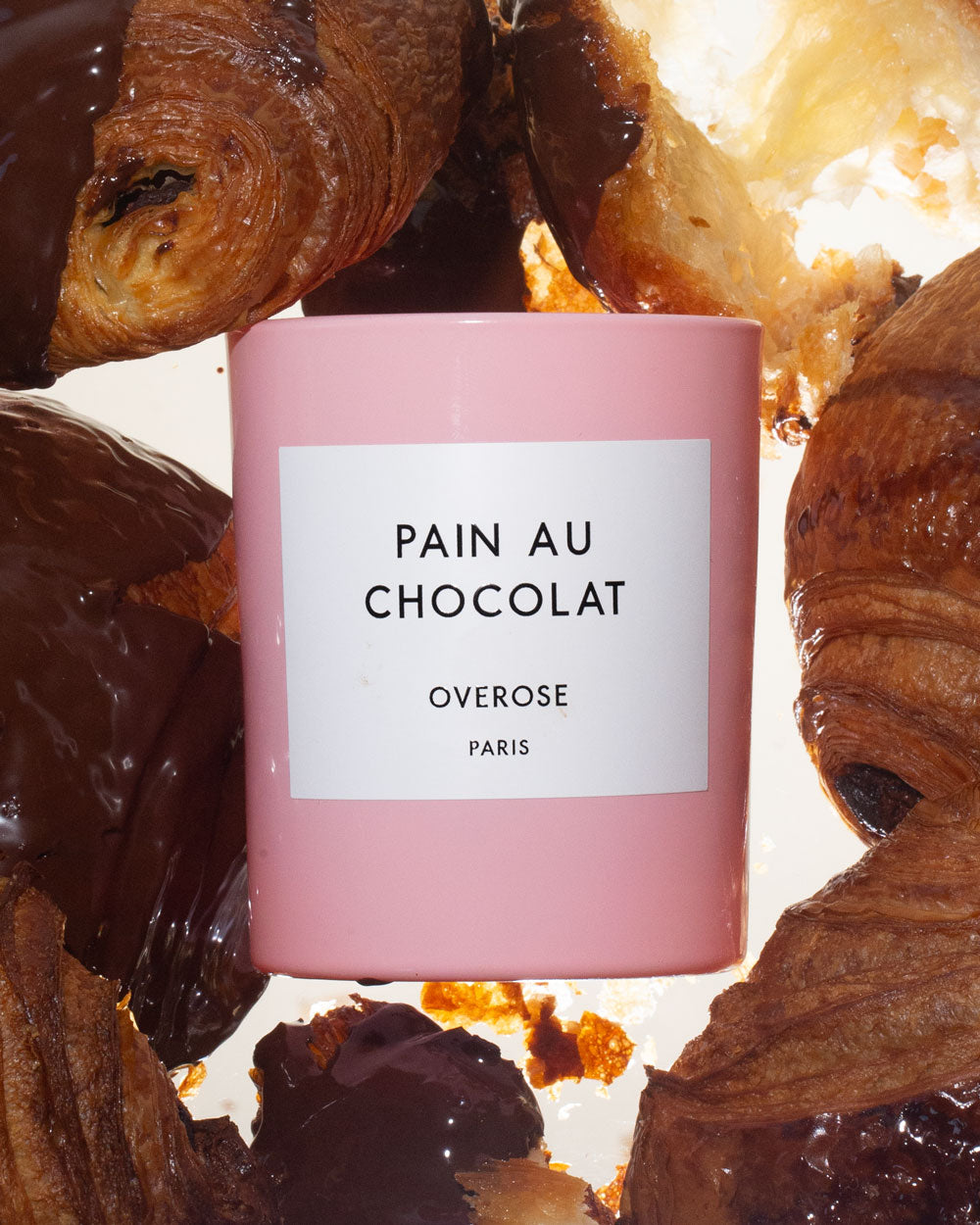 A pink candle with a label reading 'Pain au Chocolat' and 'Overose, Paris' and chocolate croissants in the background