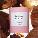 A pink candle with a label reading 'Pain au Chocolat' and 'Overose, Paris' and chocolate croissants in the background