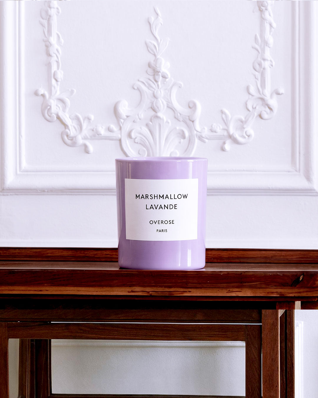 A lavender-colored candle in a cylindrical jar sits on a dark wooden table against a white wall with ornate molding.