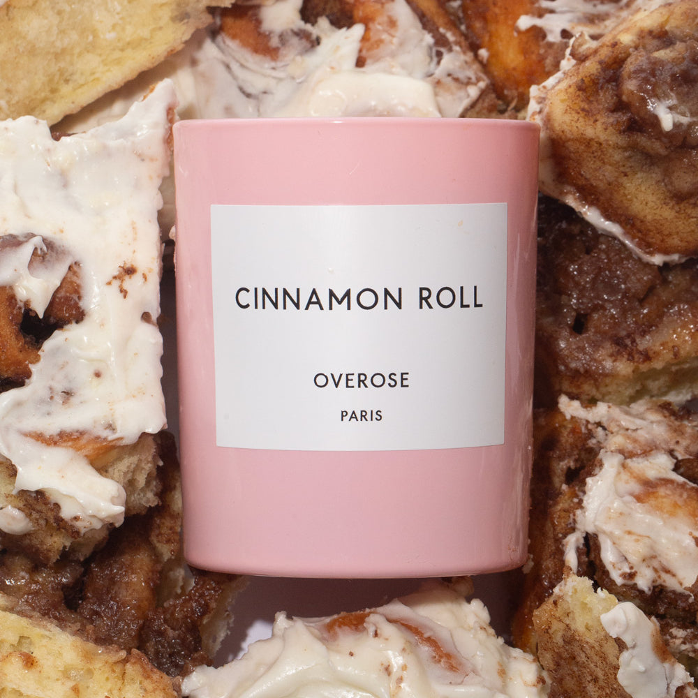 A pink cylindrical candle with the label 'Cinnamon Roll' and 'Overose Paris' on it in front of cinnamon rolls.