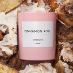 A pink cylindrical candle with the label 'Cinnamon Roll' and 'Overose Paris' on it in front of cinnamon rolls.