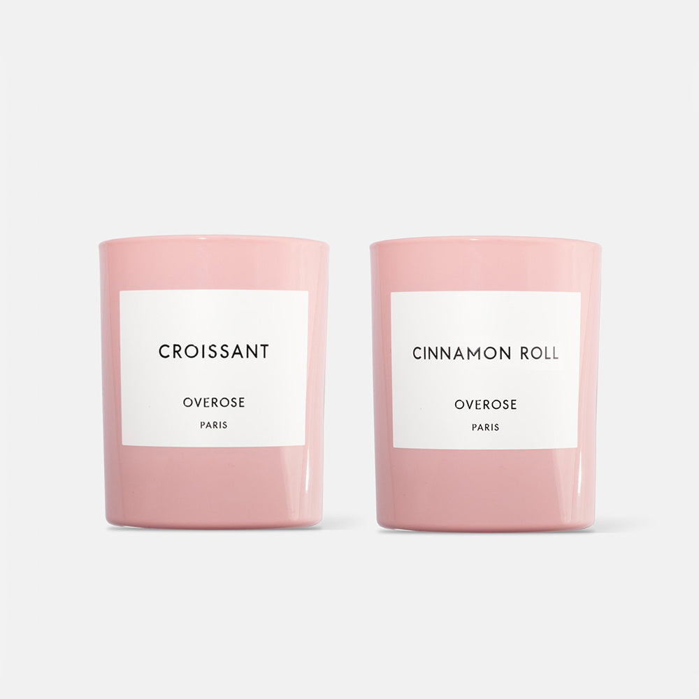 Two candles, one labeled 'Croissant' and the other 'Cinnamon Roll', each in a pink jar against a white background.